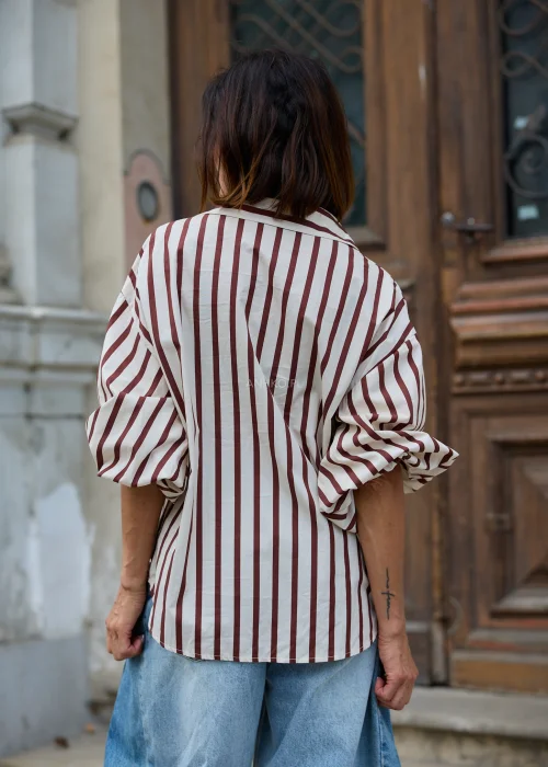 Beżowo-brązowa koszula damska oversize w pionowe paski, z klasycznym kołnierzykiem i długimi rękawami, zestawiona z jasnymi jeansami wide leg.