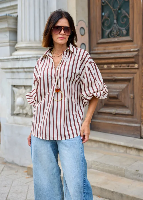 Beżowo-brązowa koszula damska oversize w pionowe paski, z klasycznym kołnierzykiem i długimi rękawami, zestawiona z jasnymi jeansami wide leg.