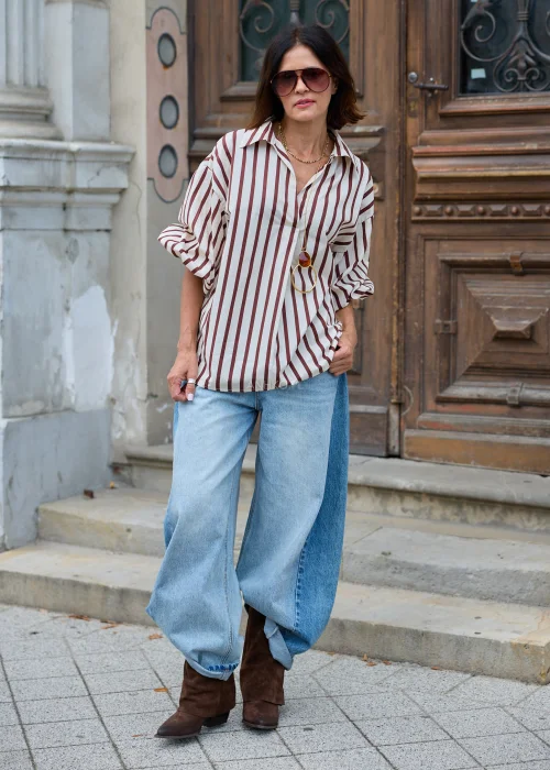 Beżowo-brązowa koszula damska oversize w pionowe paski, z klasycznym kołnierzykiem i długimi rękawami, zestawiona z jasnymi jeansami wide leg.