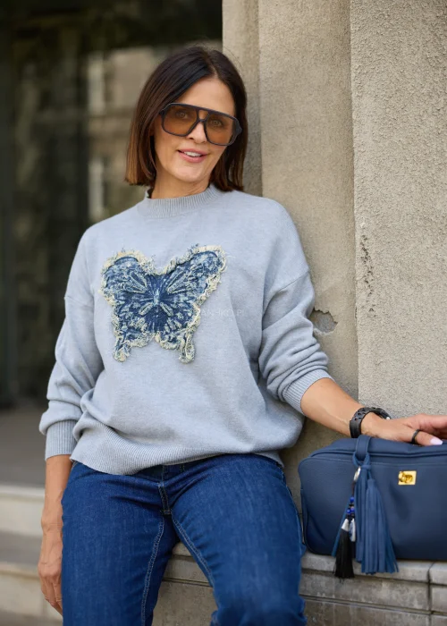 Dzianinowy sweter damski w jasnoszarym kolorze, o luźnym fasonie z długimi rękawami i dekoracyjną aplikacją motyla z denimu.