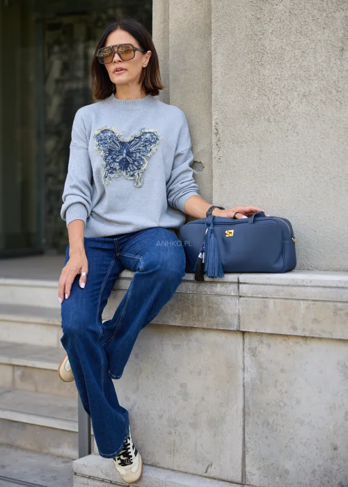 Dzianinowy sweter damski w jasnoszarym kolorze, o luźnym fasonie z długimi rękawami i dekoracyjną aplikacją motyla z denimu.