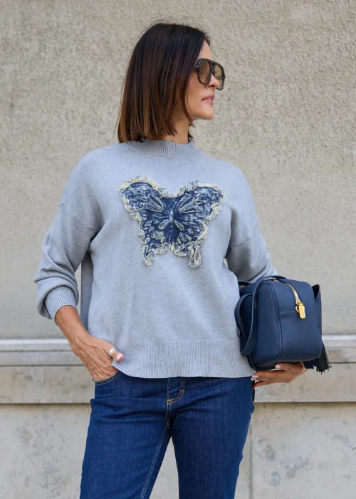 Dzianinowy sweter damski w jasnoszarym kolorze, o luźnym fasonie z długimi rękawami i dekoracyjną aplikacją motyla z denimu.