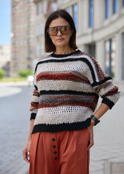 Beżowo-czarny sweter damski oversize w poziome pasy, z ażurowym splotem i rdzawo-brązowymi akcentami, zestawiony z rudymi spodniami.