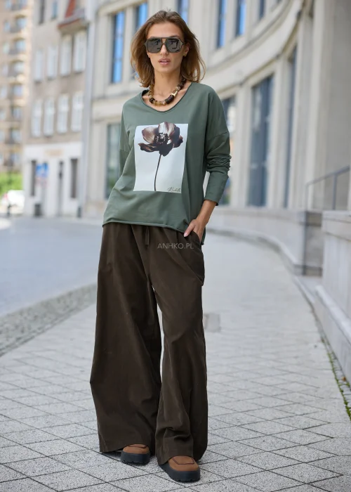 Khaki bluzka damska oversize z długim rękawem, szerokim dekoltem i dużym kwiatowym nadrukiem z przodu, zestawiona z brązowymi spodniami.