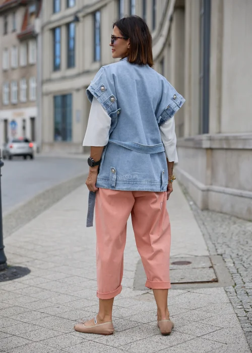 Jasnoniebieska kamizelka jeansowa damska oversize z szerokim kołnierzem, wiązanym paskiem w talii, dużymi kieszeniami i ozdobnymi metalowymi guzikami.