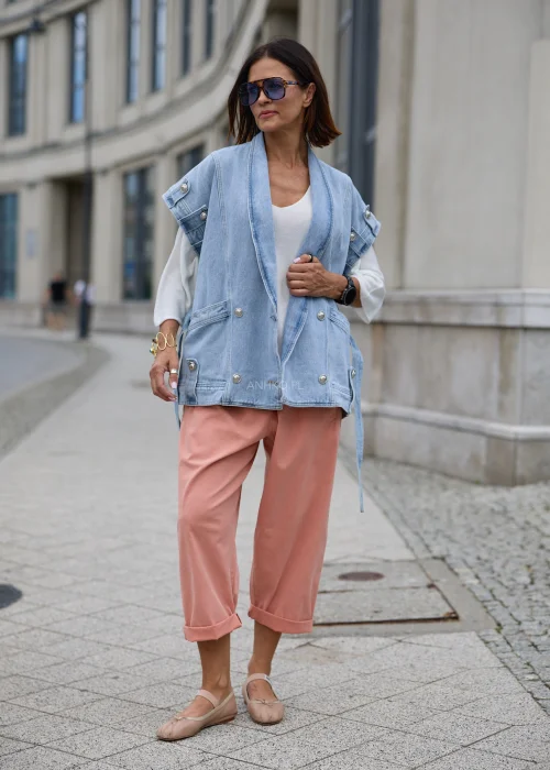 Jasnoniebieska kamizelka jeansowa damska oversize z szerokim kołnierzem, wiązanym paskiem w talii, dużymi kieszeniami i ozdobnymi metalowymi guzikami.