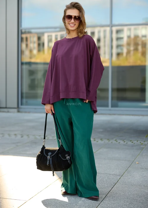 Bluzka damska oversize w kolorze śliwkowego fioletu, z luźnym, szerokim fasonem, obniżoną linią ramion, długimi rękawami, asymetrycznym warstwowym dołem oraz dekoracyjnymi wiązaniami po bokach.