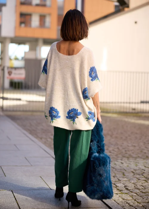 Sweter damski oversize typu ponczo z miękkiej, lekko puszystej dzianiny w kolorze kremowym, z dużym nadrukiem niebieskich kwiatów i zielonych liści, szerokim dekoltem w serek, mocno obniżoną linią ramion