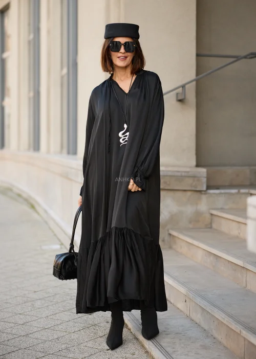 Sukienka damska maxi w kolorze czarnym, o luźnym fasonie oversize, z długimi rękawami zakończonymi marszczonymi mankietami, dekoltem w serek z wiązaniem oraz szeroką, marszczoną falbaną u dołu.