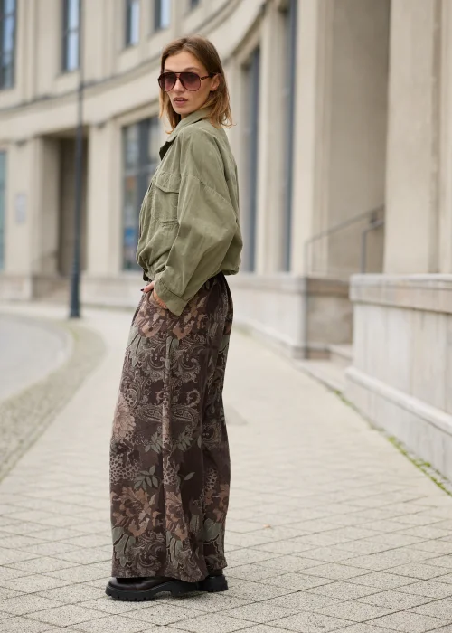 Brązowe spodnie damskie oversize z bardzo szerokimi nogawkami, kwiatowym wzorem paisley w odcieniach beżu i zieleni, elastycznym pasem, kieszeniami i ozdobnym kwiatem przy talii.