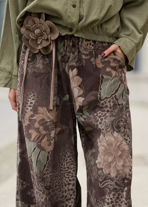 Brązowe spodnie damskie oversize z bardzo szerokimi nogawkami, kwiatowym wzorem paisley w odcieniach beżu i zieleni, elastycznym pasem, kieszeniami i ozdobnym kwiatem przy talii.