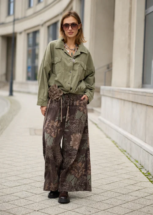 Brązowe spodnie damskie oversize z bardzo szerokimi nogawkami, kwiatowym wzorem paisley w odcieniach beżu i zieleni, elastycznym pasem, kieszeniami i ozdobnym kwiatem przy talii.