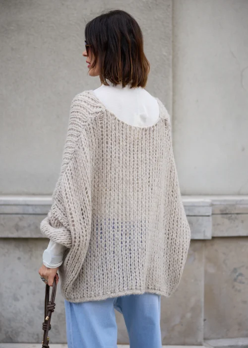 Sweter damski oversize z grubej, puszystej dzianiny o luźnym, ażurowym splocie w kolorze jasnego beżu, z okrągłym dekoltem, obniżoną linią ramion, długimi szerokimi rękawami oraz asymetrycznym, łukowato wyciętym dołem.