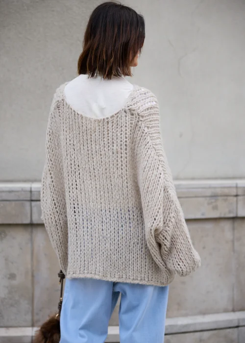 Sweter damski oversize z grubej, puszystej dzianiny o luźnym, ażurowym splocie w kolorze jasnego beżu, z okrągłym dekoltem, obniżoną linią ramion, długimi szerokimi rękawami oraz asymetrycznym, łukowato wyciętym dołem.