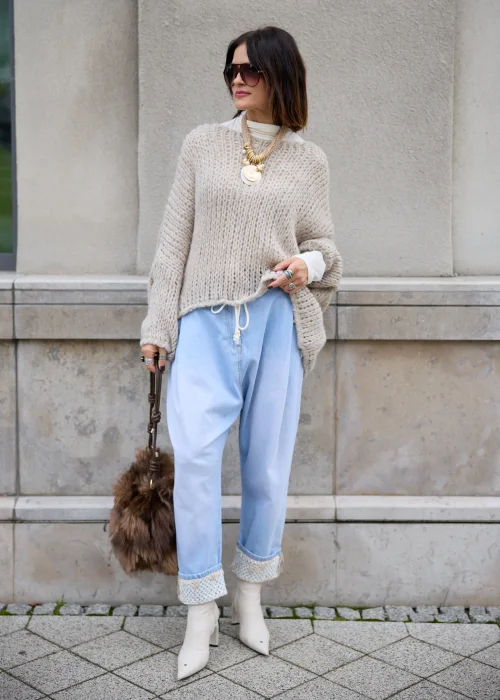 Sweter damski oversize z grubej, puszystej dzianiny o luźnym, ażurowym splocie w kolorze jasnego beżu, z okrągłym dekoltem, obniżoną linią ramion, długimi szerokimi rękawami oraz asymetrycznym, łukowato wyciętym dołem.