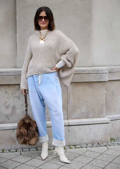 Sweter damski oversize z grubej, puszystej dzianiny o luźnym, ażurowym splocie w kolorze jasnego beżu, z okrągłym dekoltem, obniżoną linią ramion, długimi szerokimi rękawami oraz asymetrycznym, łukowato wyciętym dołem.