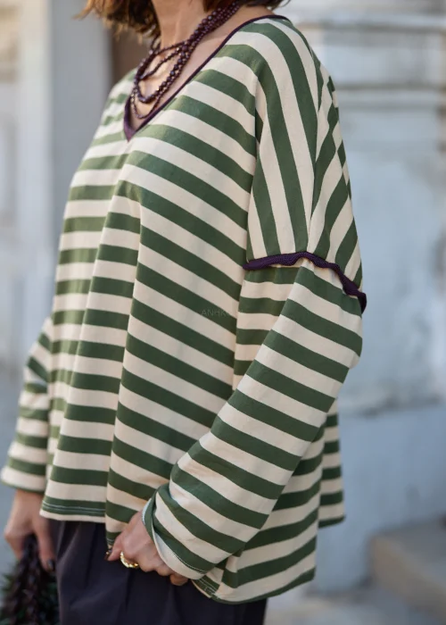 Bluzka damska oversize w szerokie poziome paski w kolorze oliwkowej zieleni i kremowego beżu, z dekoltem w serek, kontrastowym fioletowym wykończeniem, obniżoną linią ramion, długimi szerokimi rękawami oraz luźnym fasonem.