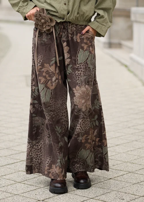 Brązowe spodnie damskie oversize z bardzo szerokimi nogawkami, kwiatowym wzorem paisley w odcieniach beżu i zieleni, elastycznym pasem, kieszeniami i ozdobnym kwiatem przy talii.