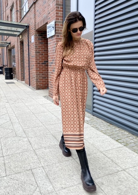 Wzorzysta sukienka midi z wiskozy w retro print w odcieniach brązu, z marszczonym dekoltem i gumką w pasie. Stylizacja z jasnymi kowbojkami i czarną torebką podkreśla modowy charakter modelu.
