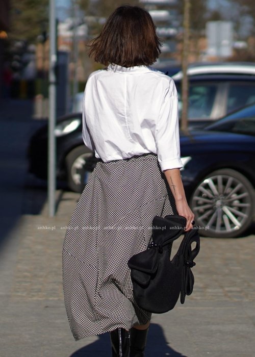 Biała koszula damska oversize z kołnierzykiem i przeszyciem poniżej biustu, zestawiona z czarno-białą spódnicą w geometryczny wzór w miejskiej stylizacji.