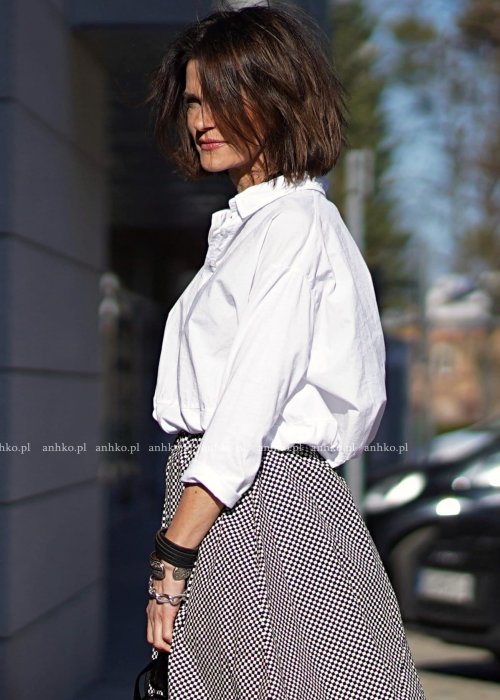 Biała koszula damska oversize z kołnierzykiem i przeszyciem poniżej biustu, zestawiona z czarno-białą spódnicą w geometryczny wzór w miejskiej stylizacji.