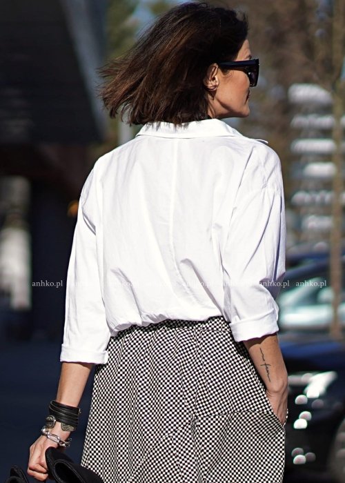 Biała koszula damska oversize z kołnierzykiem i przeszyciem poniżej biustu, zestawiona z czarno-białą spódnicą w geometryczny wzór w miejskiej stylizacji.