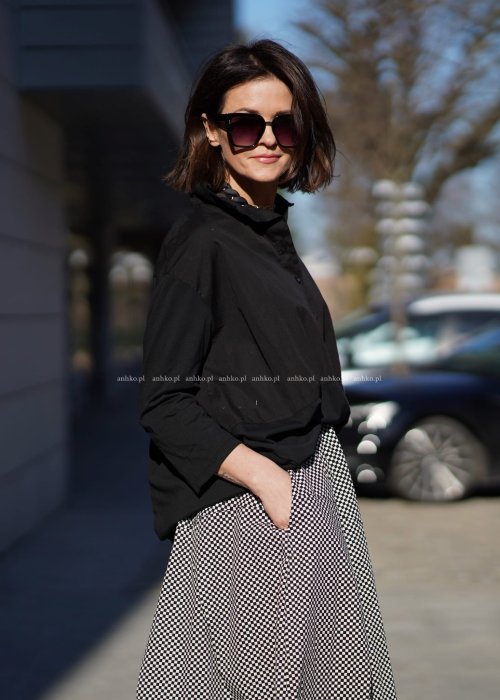 Czarna koszula damska oversize z kołnierzykiem i przeszyciem poniżej biustu, zestawiona z czarno-białą spódnicą w geometryczny wzór w miejskiej stylizacji.