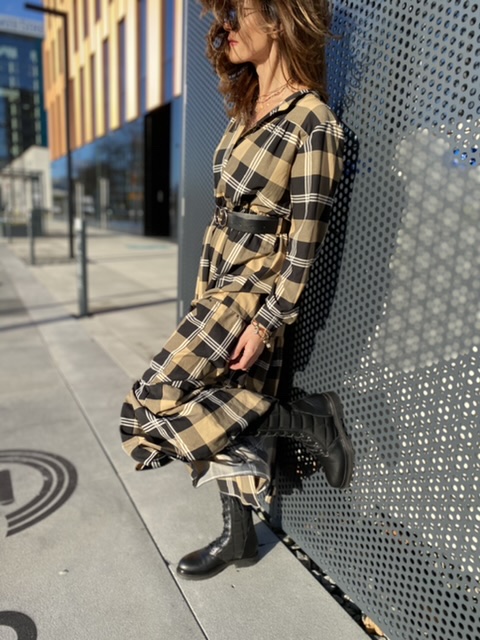 Sukienka maxi w beżowo-czarną kratę z paskiem w talii, o luźnym kroju i warstwowym dole, stylizowana z ciężkimi botkami i dużą torebką.