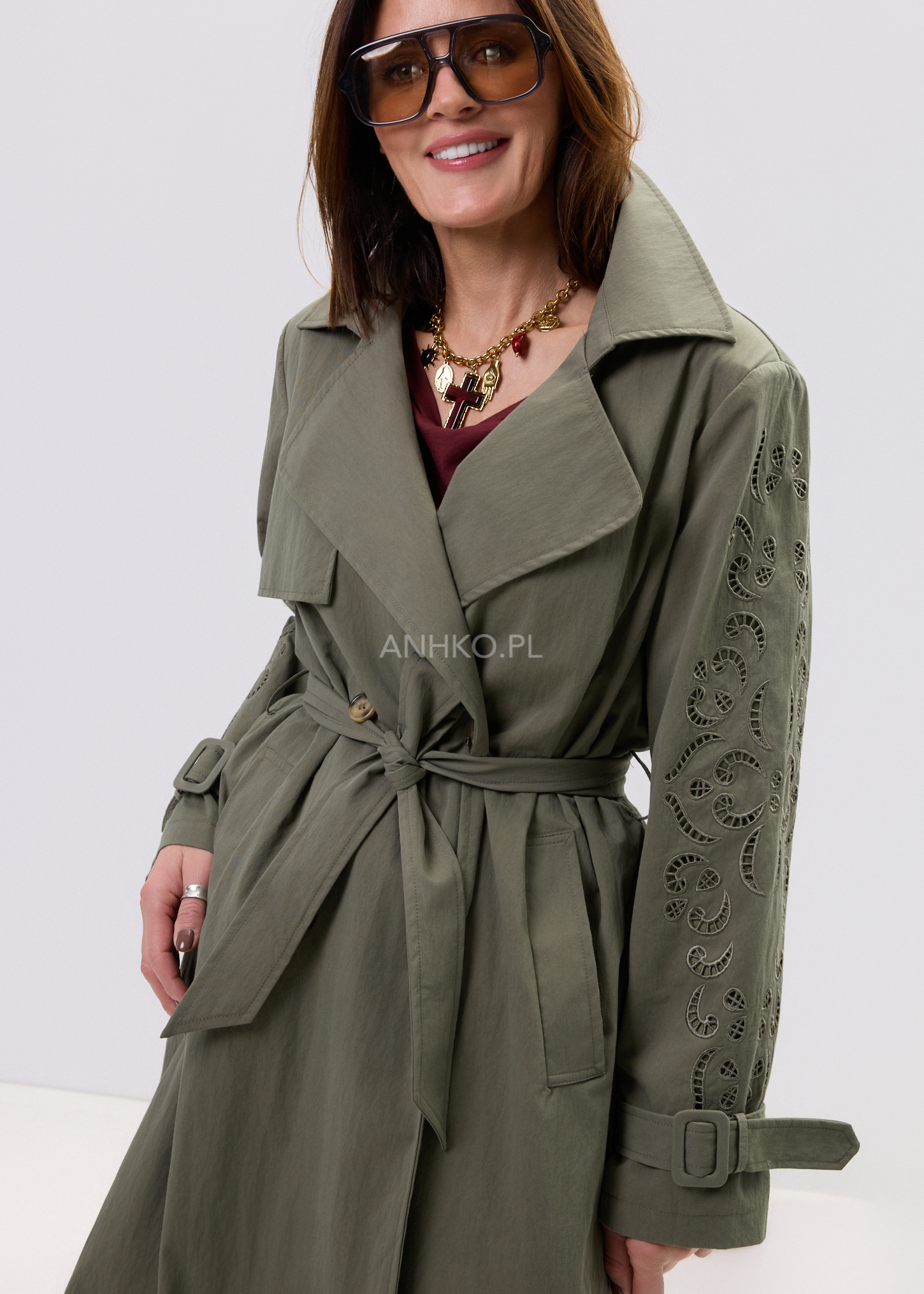 Damski trencz khaki midi z paskiem w talii i szerokimi klapami, model z ozdobnymi ażurowymi zdobieniami na rękawach w eleganckiej stylizacji.