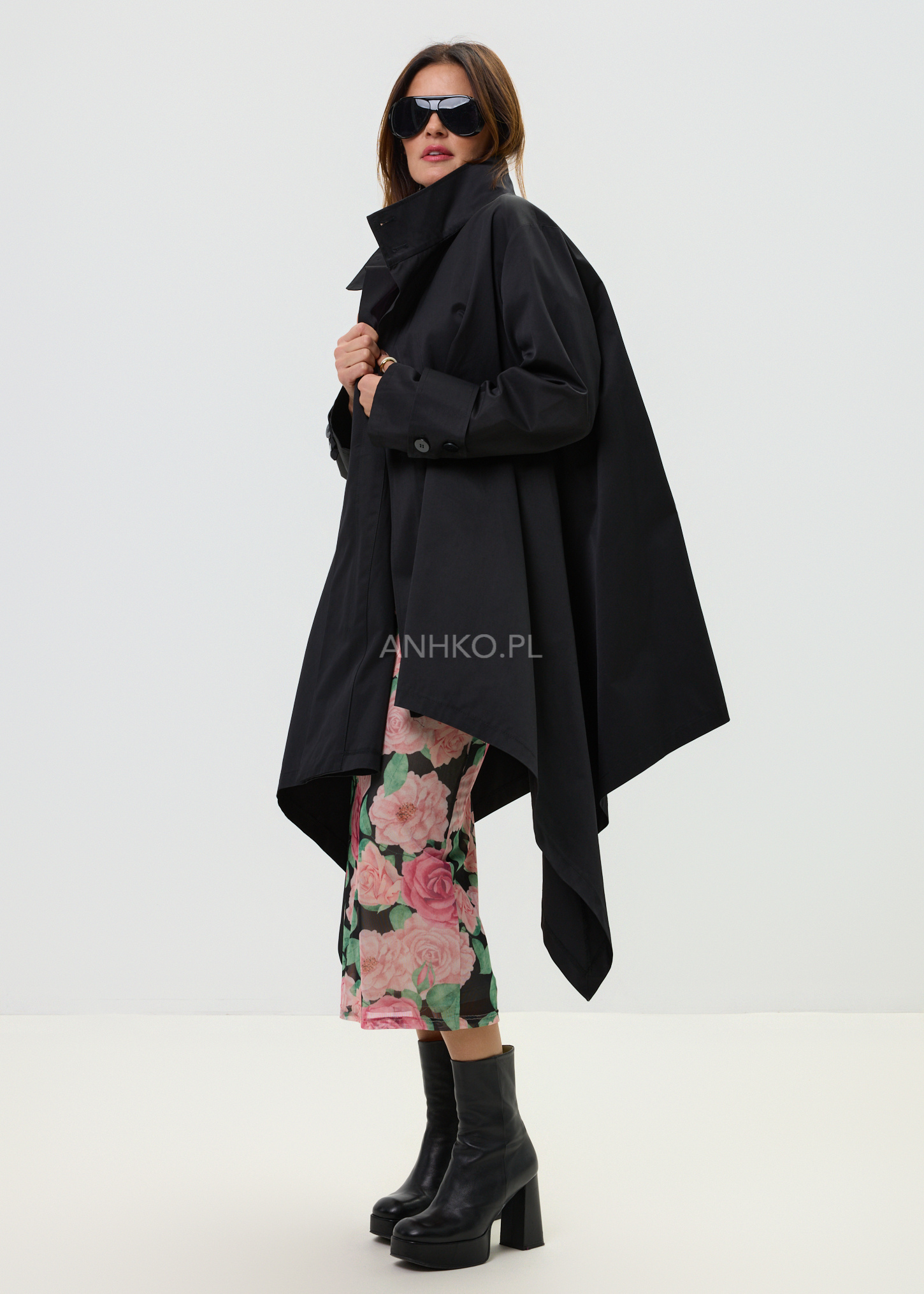 Czarny damski płaszcz o asymetrycznym kroju z wysokim kołnierzem, elegancka stylizacja z kwiatową spódnicą midi i czarnymi botkami.