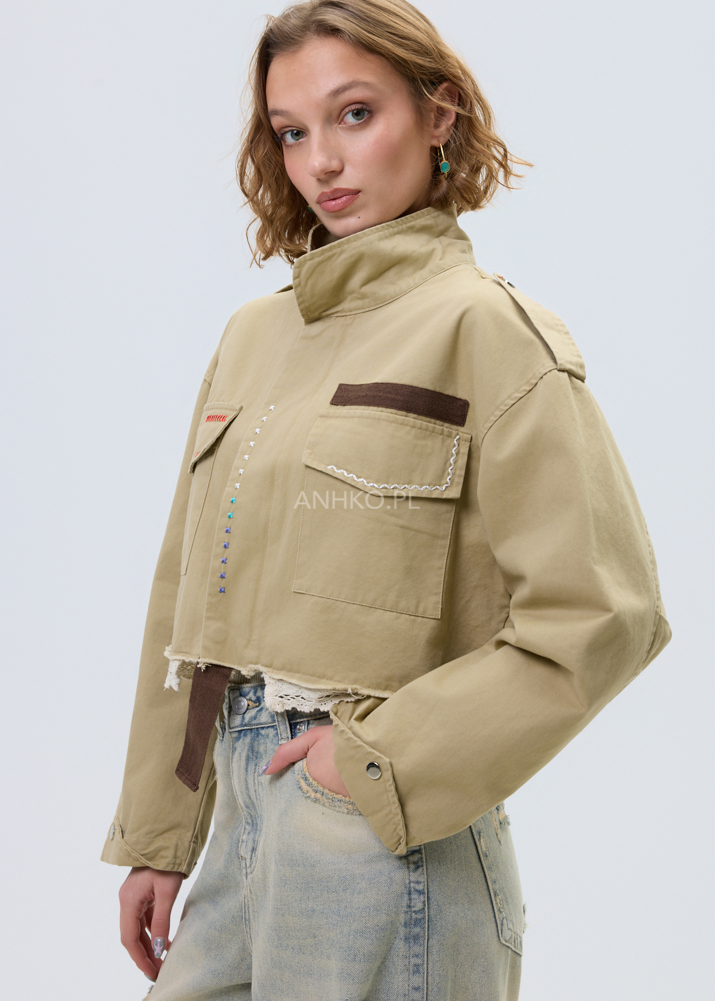 Krótka damska kurtka khaki z wysoką stójką, kieszeniami z przodu i ozdobnymi przeszyciami, stylizacja z jasnymi jeansami w casualowym stylu.