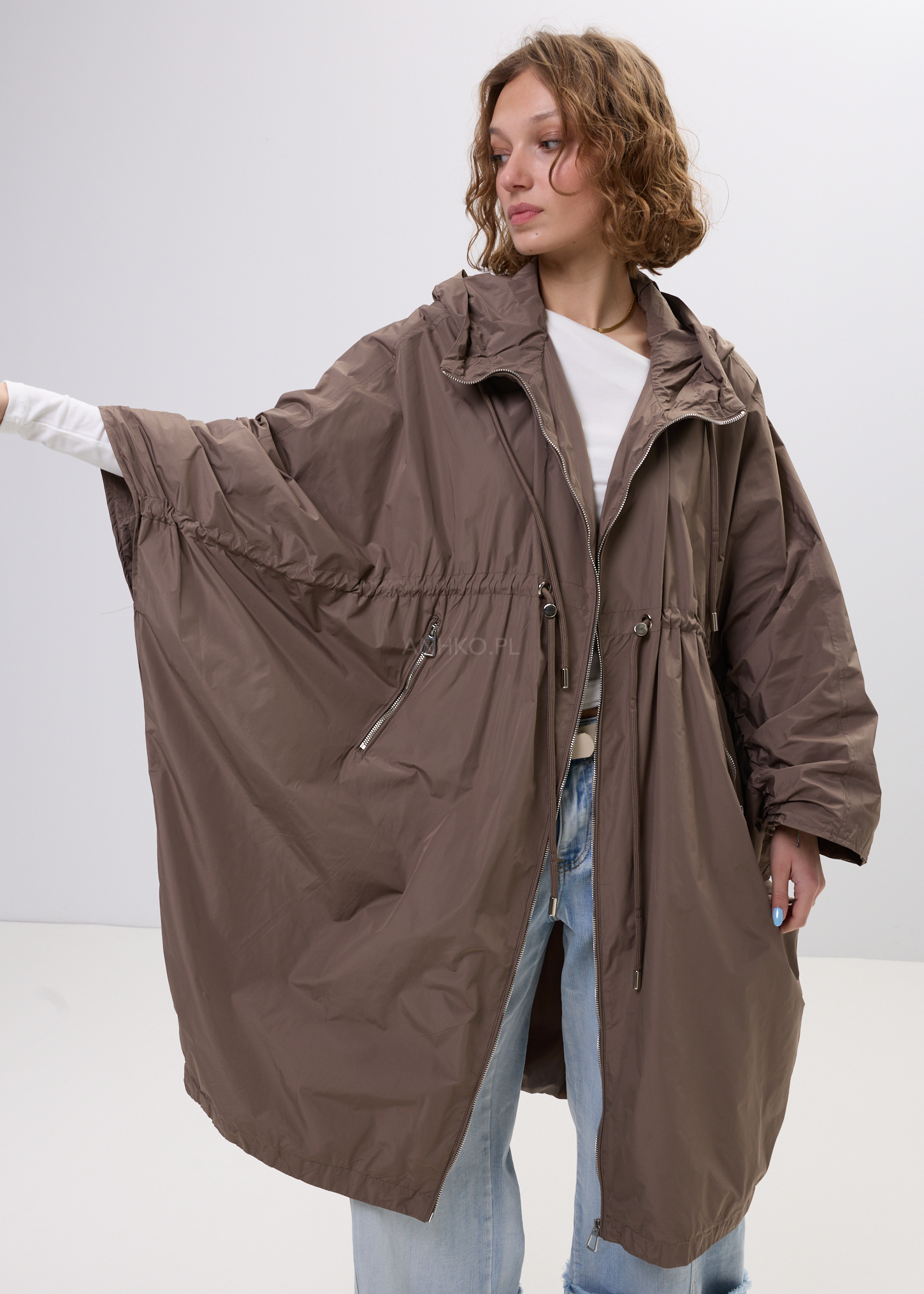 Damski płaszcz przeciwdeszczowy o luźnym kroju w brązowym kolorze z kapturem, zapinany na zamek z regulacją w talii i kieszeniami na zamek.