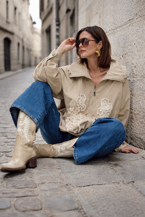 Beżowa damska kurtka oversize zapinana na zamek z wysoką stójką i ozdobnymi aplikacjami z przodu, stylizacja z jeansami i kowbojkami w miejskim stylu.
